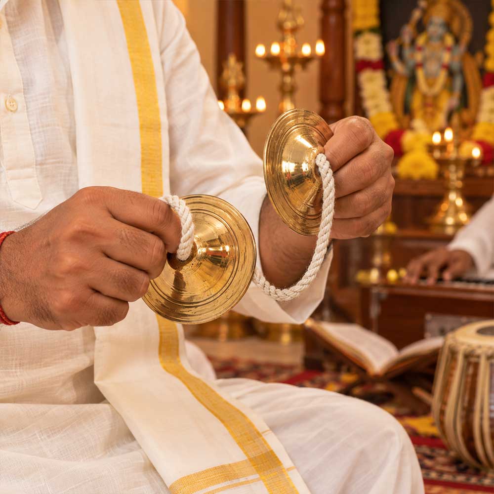 A close-up of a person in a white traditional outfit with a gold stripe holding two small brass cymbals connected by a white rope, with a temple interior and lit brass lamps in the background.